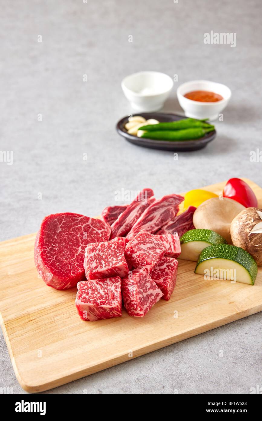 Premium koreanische Hanwoo-Wagyu-Rindfleischstücke mit starker Marmorierung, die auf einem Holzschneidebrett mit frischem Gemüse inklusive grüner Paprika und Pilz präsentiert werden Stockfoto