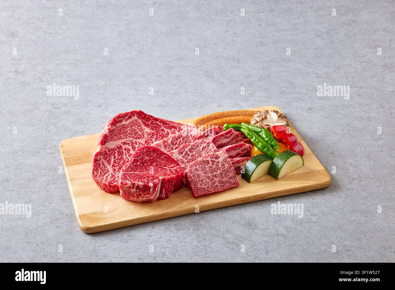 Premium koreanische Hanwoo-Wagyu-Rindfleischstücke mit starker Marmorierung, die auf einem Holzschneidebrett mit frischem Gemüse inklusive grüner Paprika und Pilz präsentiert werden Stockfoto