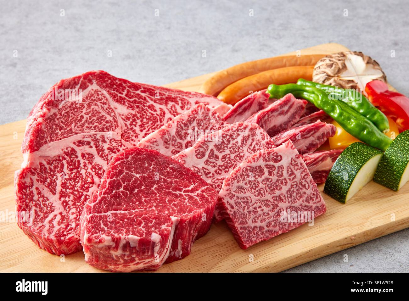 Premium koreanische Hanwoo-Wagyu-Rindfleischstücke mit starker Marmorierung, die auf einem Holzschneidebrett mit frischem Gemüse inklusive grüner Paprika und Pilz präsentiert werden Stockfoto