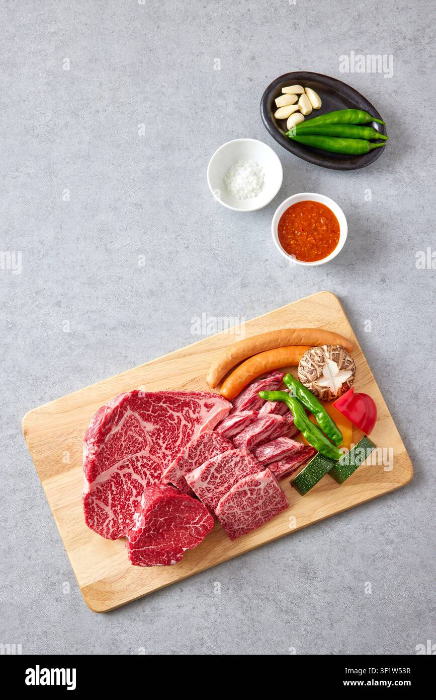 Premium koreanische Hanwoo-Wagyu-Rindfleischstücke mit starker Marmorierung, die auf einem Holzschneidebrett mit frischem Gemüse inklusive grüner Paprika und Pilz präsentiert werden Stockfoto