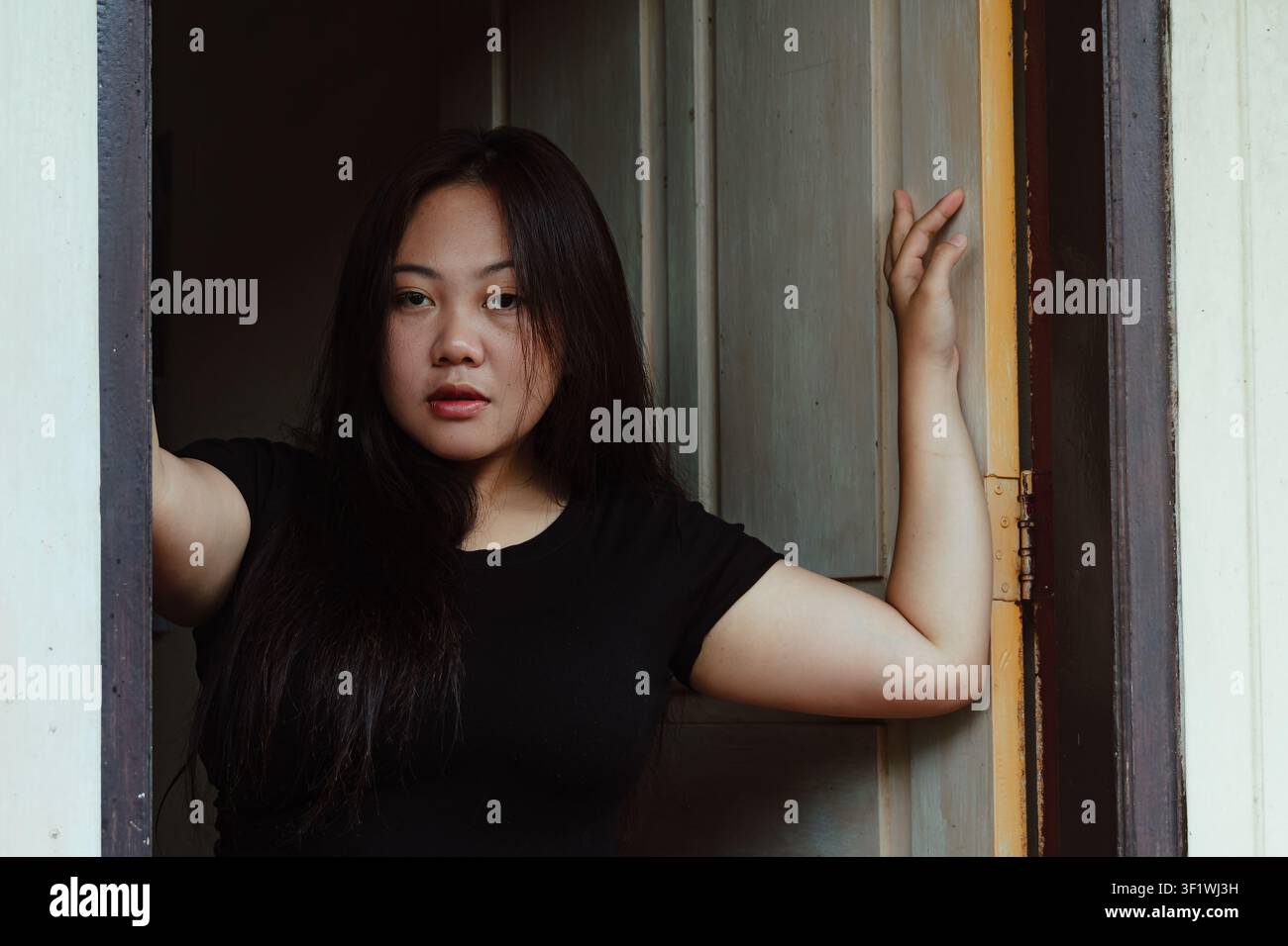 Schöne asiatische Frau steht in einem alten hölzernen Türrahmen und schaut direkt in die Kamera mit einem selbstbewussten Ausdruck. Sie trägt ein schlichtes schwarzes T-Shirt. Stockfoto