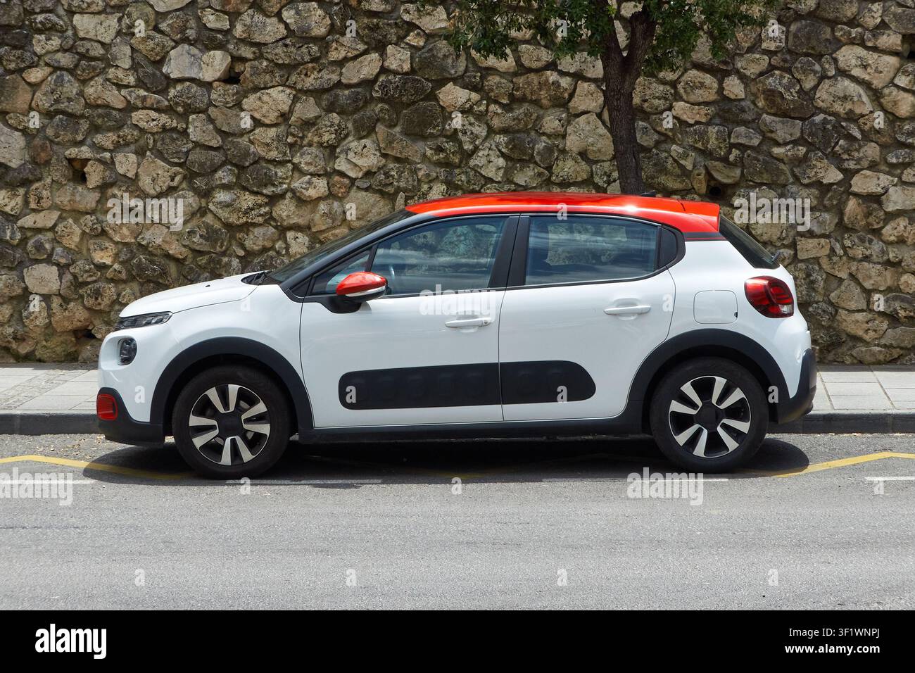 Weißer und orangefarbener Citroën C3, der an der Straße vor einer Steinmauer im Hintergrund geparkt ist, Seitenansicht. Stockfoto