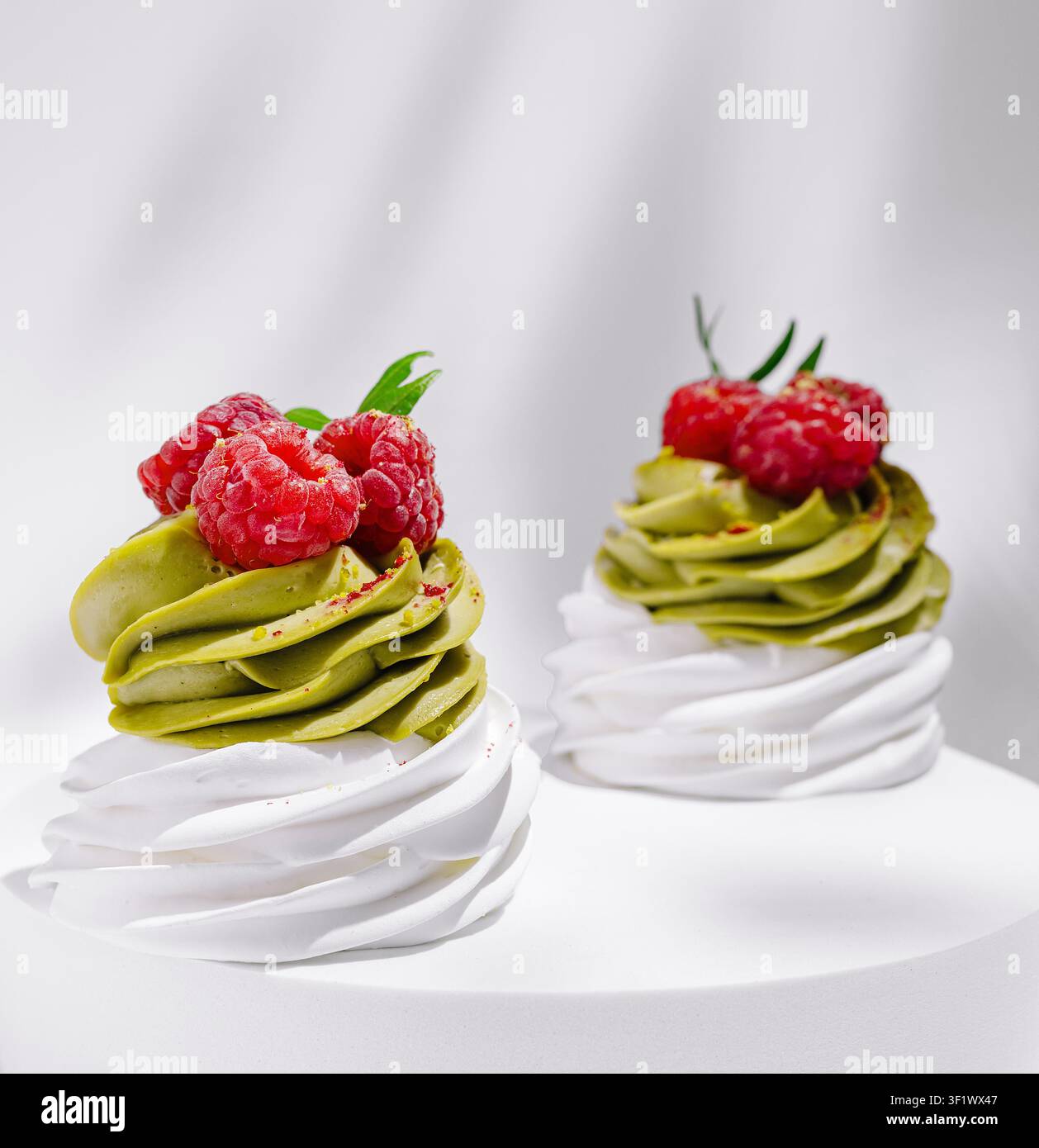 Zwei elegante Gebäckstücke mit wirbelnder grüner Matcha-Creme, gekrönt mit reifen Himbeeren, sitzen auf einem einfachen weißen Ständer. Die süßen Leckereien zeigen eine perfekte Mischung aus Aromen und Farben. Stockfoto