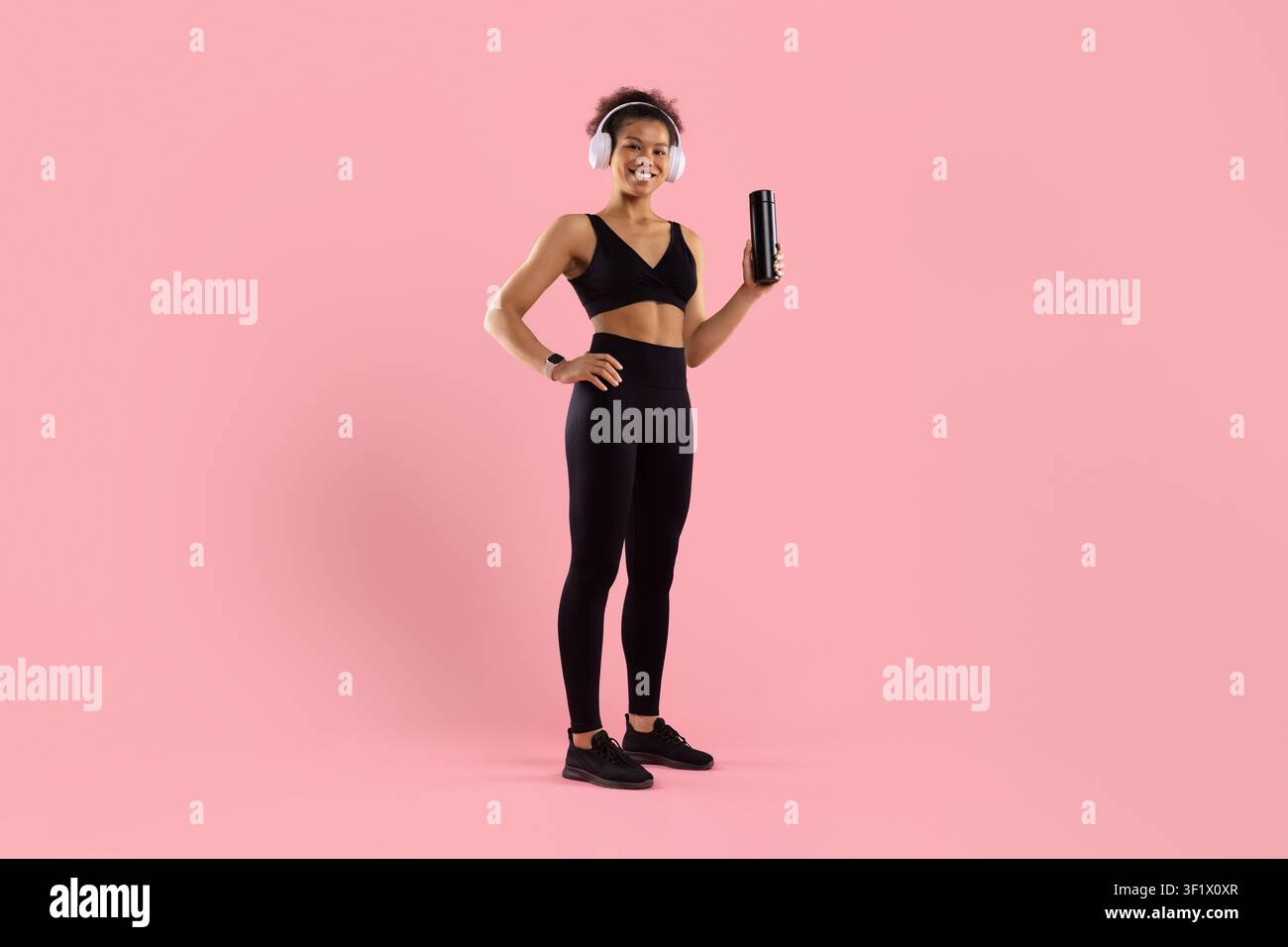 Vollständige Körperdarstellung einer glücklichen schwarzen Frau, die Kopfhörer trägt und eine Wasserflasche hält Stockfoto