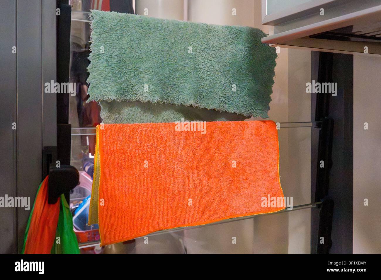 Zwei farbenfrohe Putztücher hängen an einem Glastürgriff. Eines ist grün, das andere leuchtend orange. Sie wirken weich und saugfähig, geeignet für die Reinigung Stockfoto