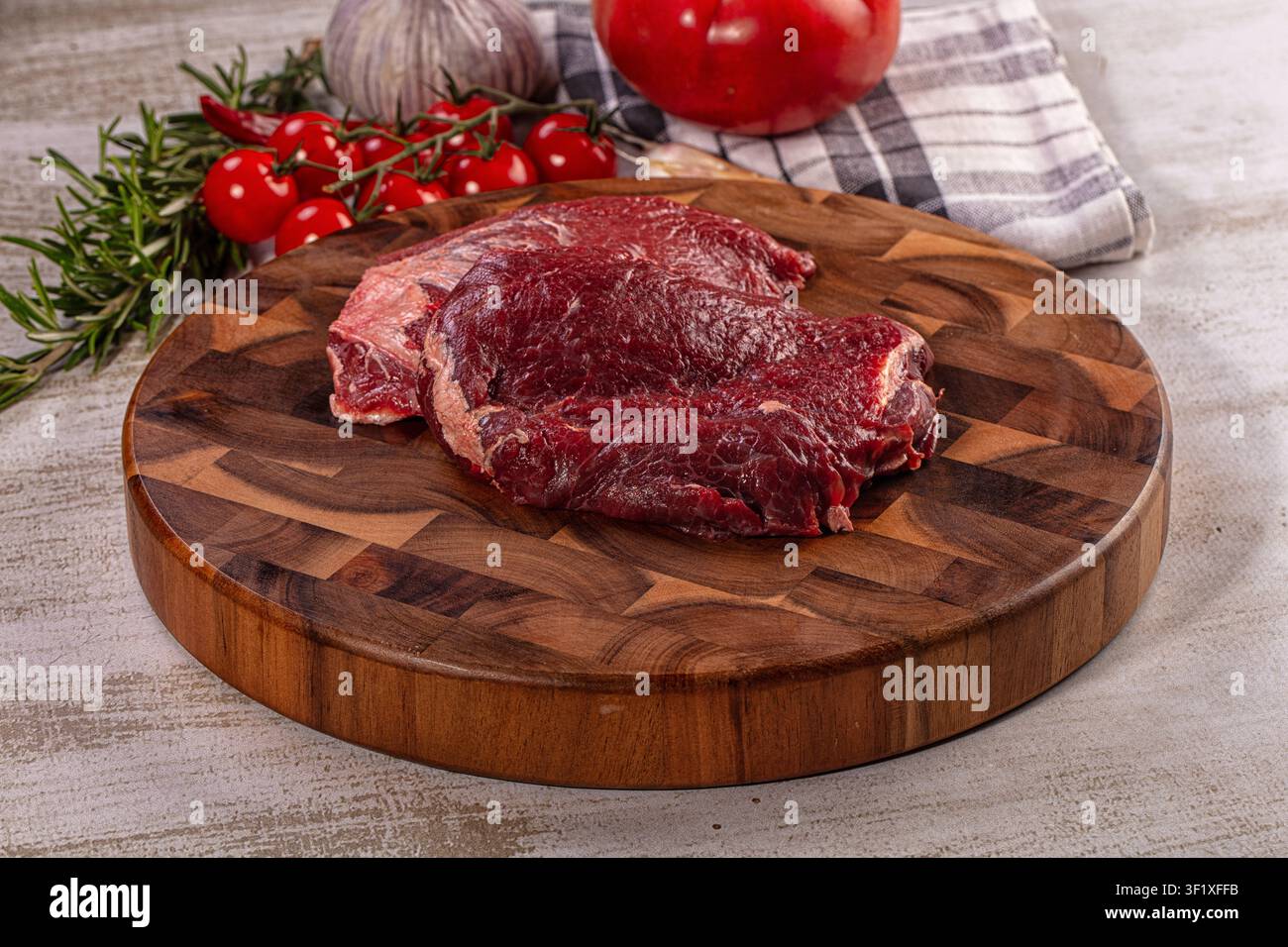 Rohes Rindfleisch-Wangensteak auf rundem hölzernem Schneidebrett. Frische Kirschtomaten, Knoblauch, Rosmarin und roter Pfeffer in der Nähe. Zutaten für Gourmet-Kochen Stockfoto