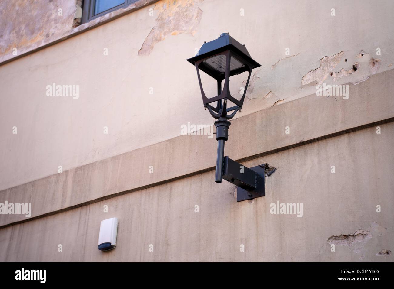 schwarze Metalllaterne auf einer Befestigungsklammer, die an einer verwitterten beigen Stuckfassade mit abblätterndem Putz und subtilen Schatten angebracht ist, die eine Ruhe ausstrahlt Stockfoto