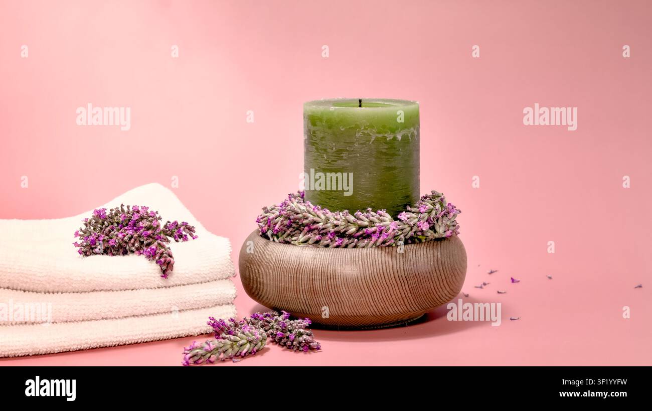 Weiße Handtücher, eine hohe grüne Kerze und frische Lavendelstiele in und um eine strukturierte Holzschale schaffen eine beruhigende Spa-Atmosphäre. Stockfoto