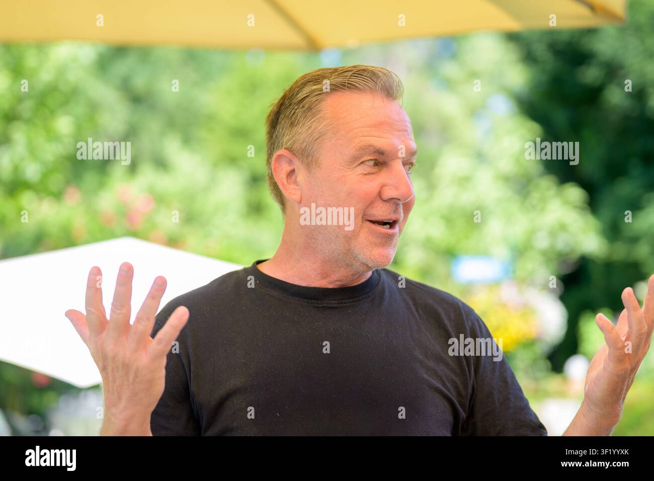 Ein Mann mittleren Alters gestikuliert ausdrucksvoll, während er im Freien spricht, und fängt Kommunikation, Diskussion und authentische menschliche Interaktion ein. Stockfoto