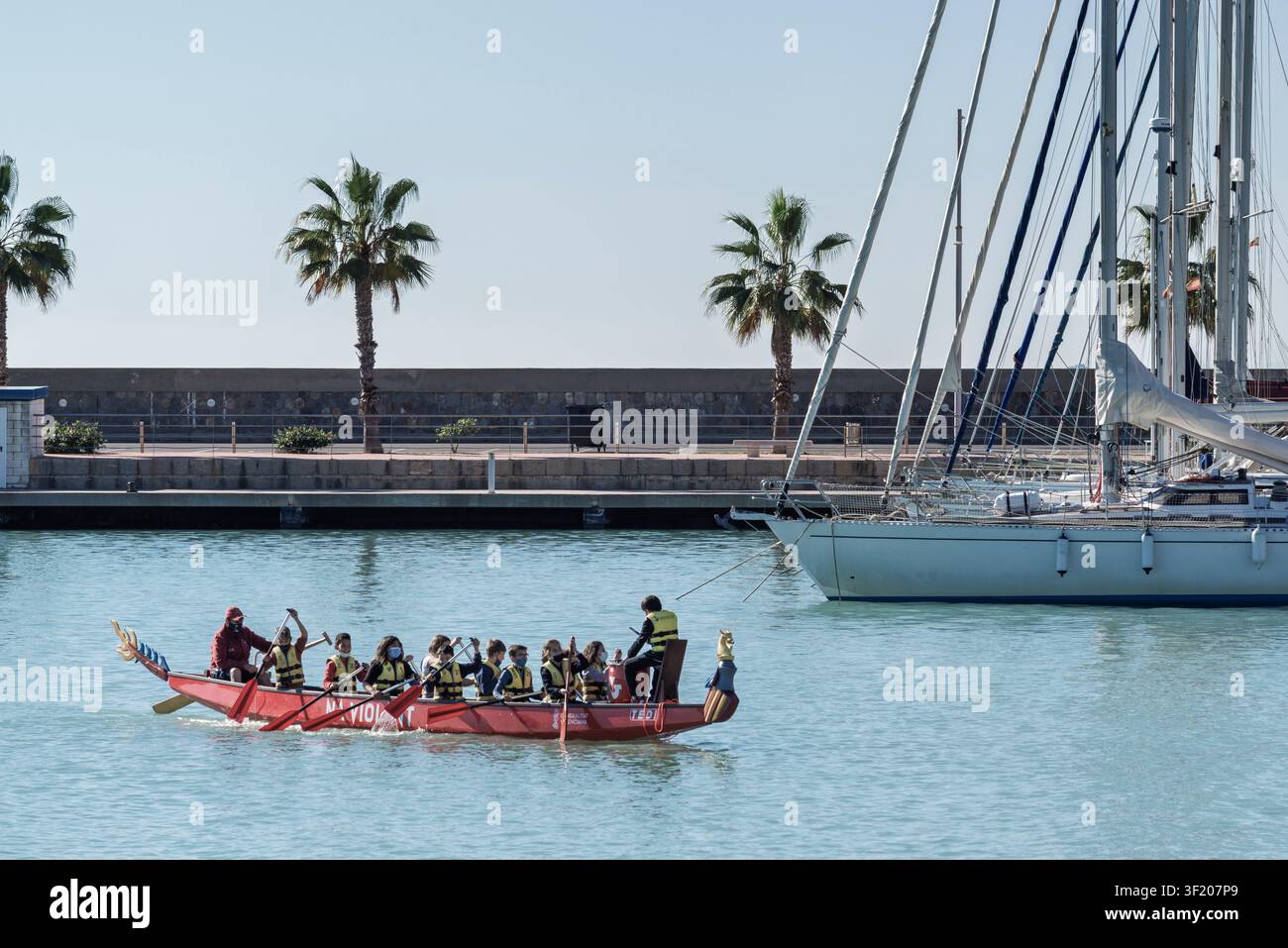 Kinder rudern ein rotes Drachenboot im Hafen von Burriana, Borriana, Castello Costa del Azahar, Spanien. Segelboote und Palmen rahmen die sonnige nautische Szene ein Stockfoto