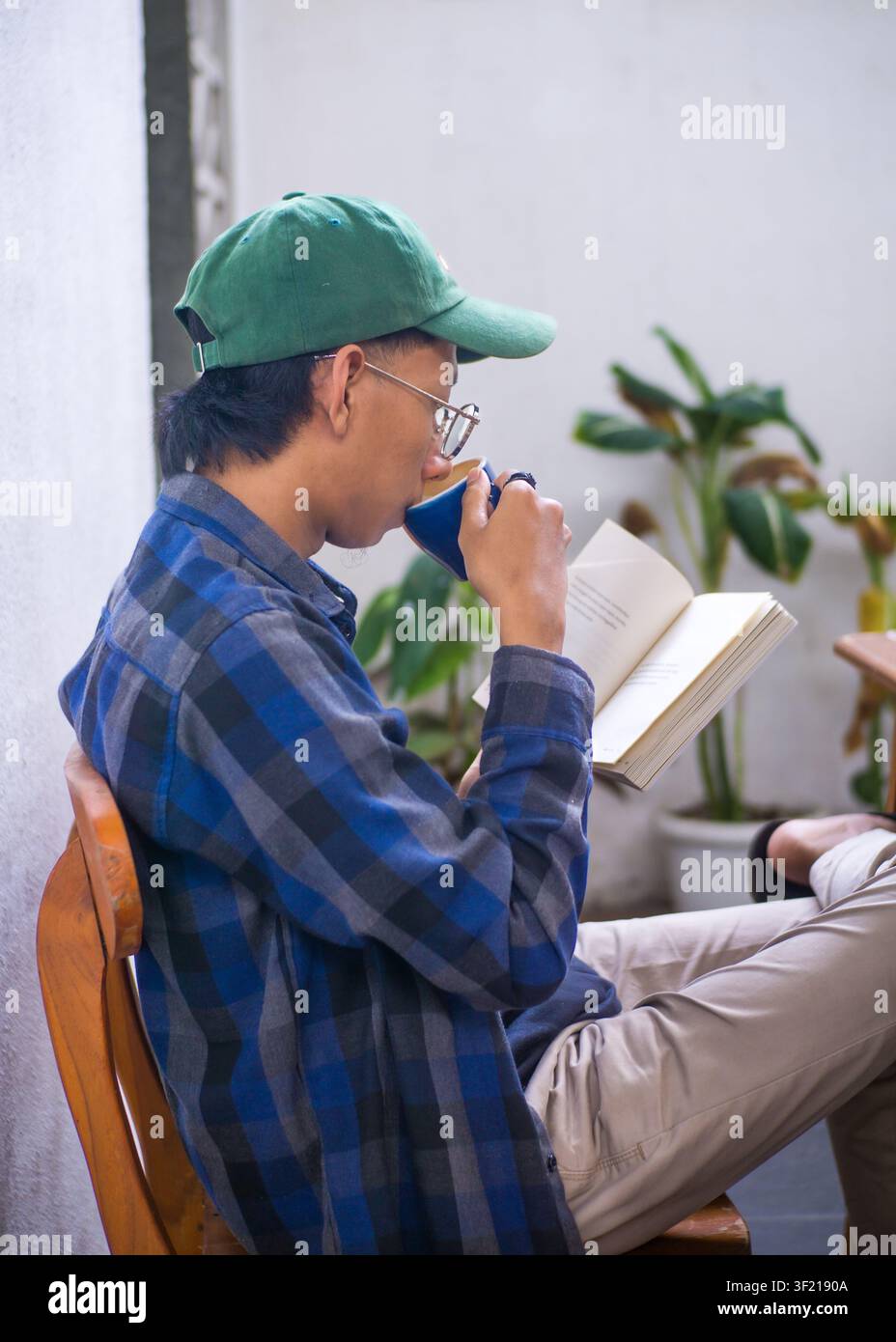 Junger asiatischer Mann liest ein Buch und genießt dabei eine Tasse Kaffee in einem gemütlichen Café, authentisches Lifestyle-Porträt. Stockfoto