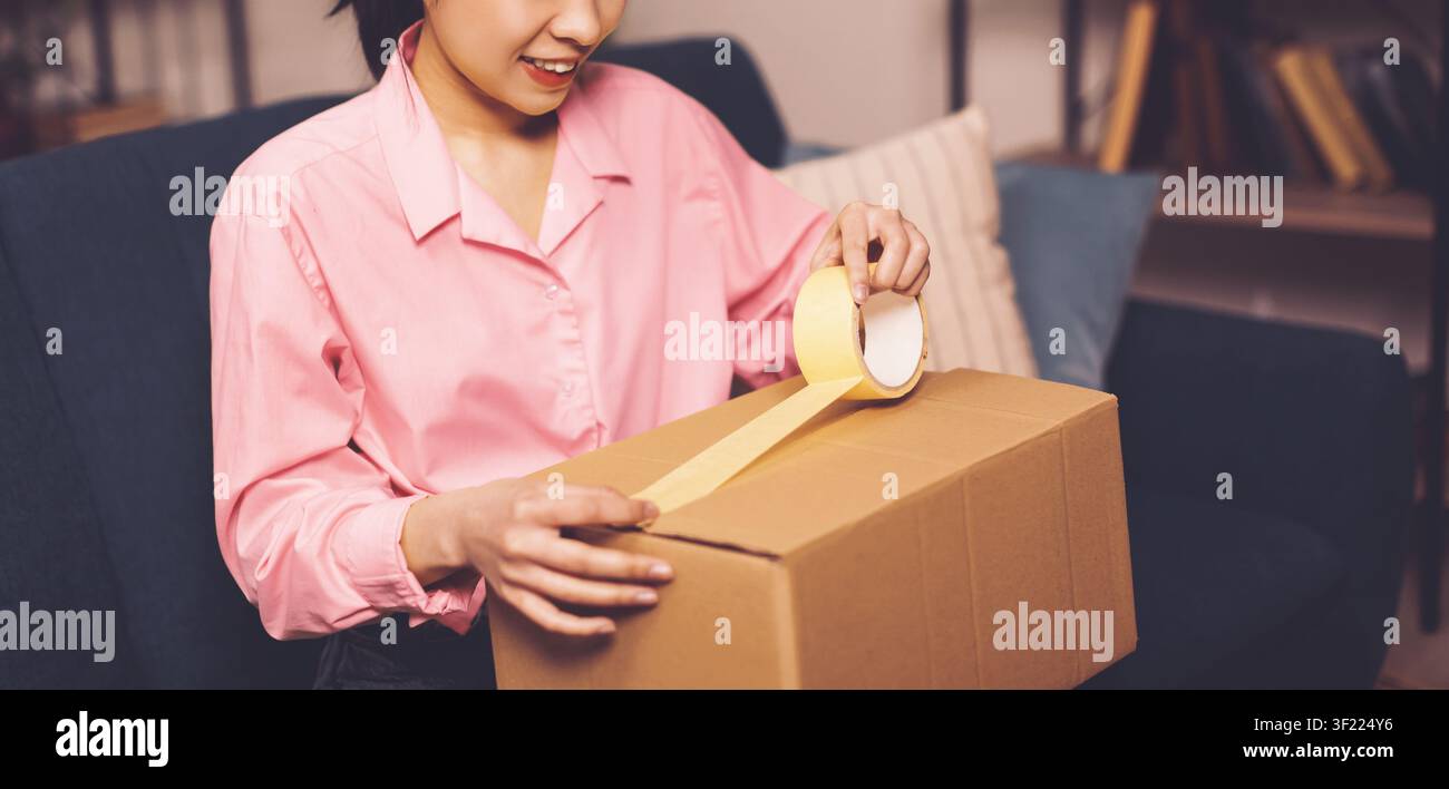 Junge asiatische Frau packt eine Kiste zu Hause mit Klebeband, Lieferkonzept Stockfoto