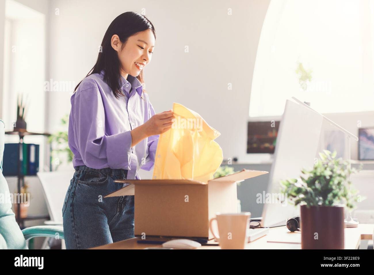Zufriedene asiatische Frau, die eine Lieferbox in einem hellen modernen Büro auspackt, Begeisterungskonzept Stockfoto