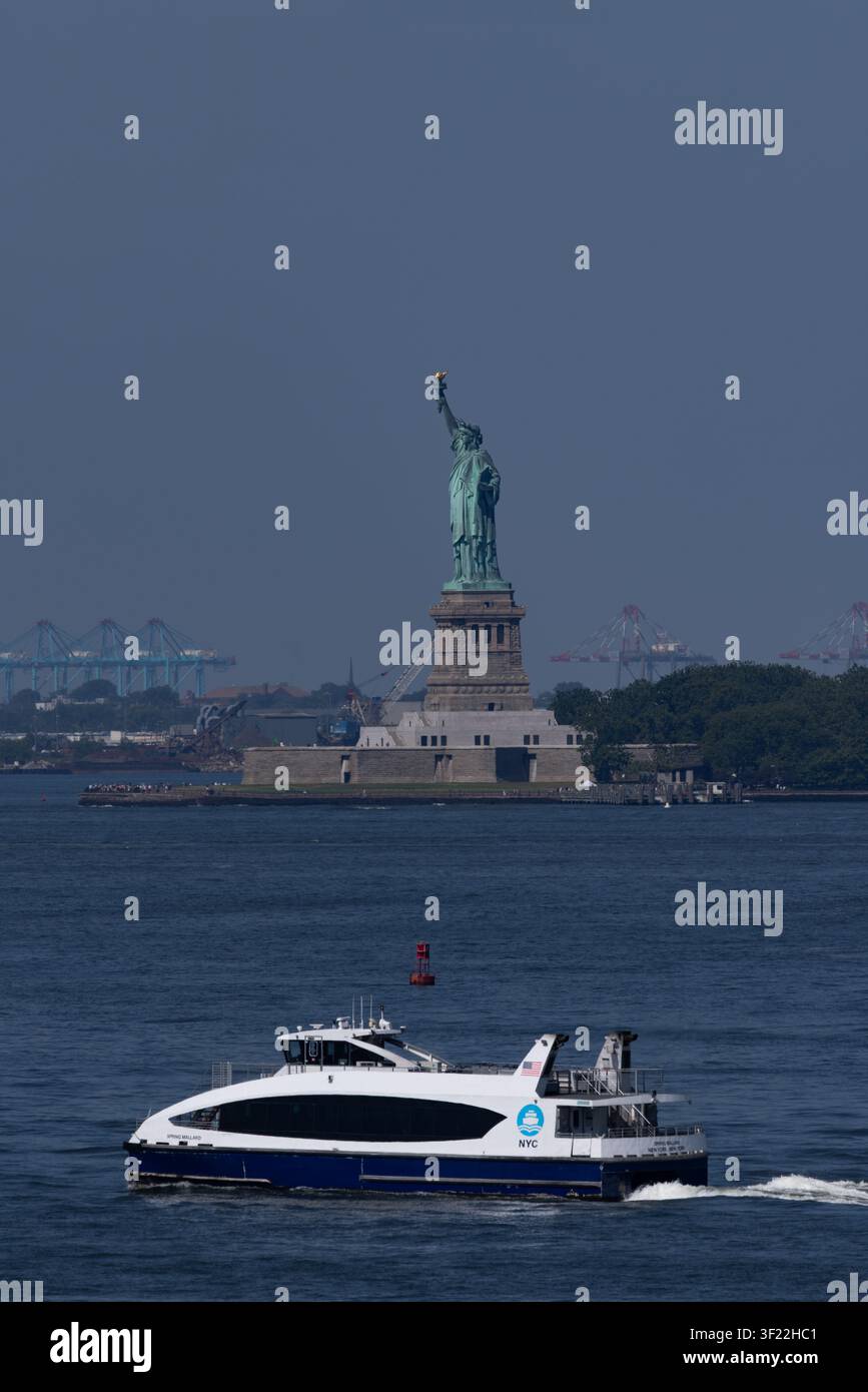 BROOKLYN, NY - 3. JULI 2026: NYC-Fährenboot fährt vor dem Statue of Liberty National Monument im New York Harbor vorbei, Blick von Brooklyn Stockfoto