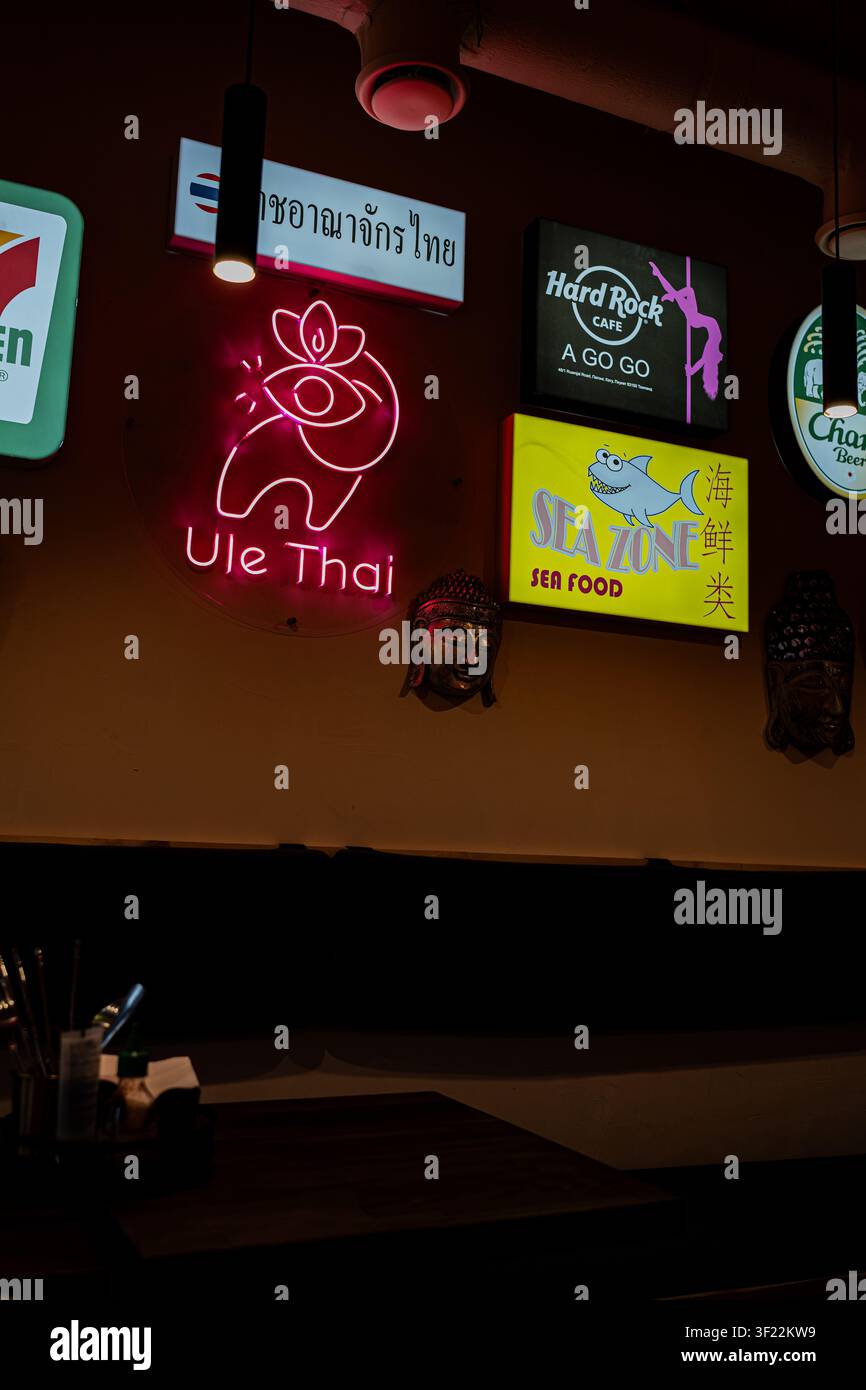 Schwach beleuchtetes Restaurantinnere mit Neonreklamen inklusive Ule Thai, Hard Rock Cafe und Sea Zone Seafood. Asiatische Baratmosphäre mit Neonlichtern. Stockfoto