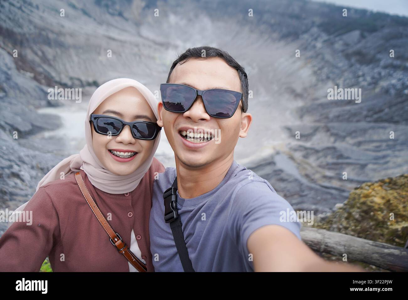 Lächelndes asiatisches Paar mit Sonnenbrille, das ein fröhliches Selfie mit einem malerischen Vulkankrater im Hintergrund macht. Stockfoto
