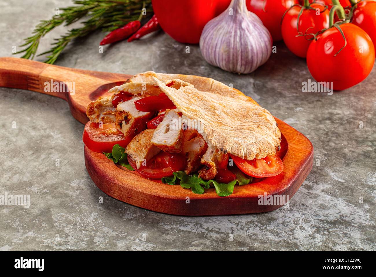 Gegrilltes Hühnchen-Pita-Sandwich mit frischen Tomatenscheiben und Salat auf rustikalem Holzbrett. Im Hintergrund sind Knoblauch, Rosmarin, Chilischoten und Weintrauben zu sehen Stockfoto