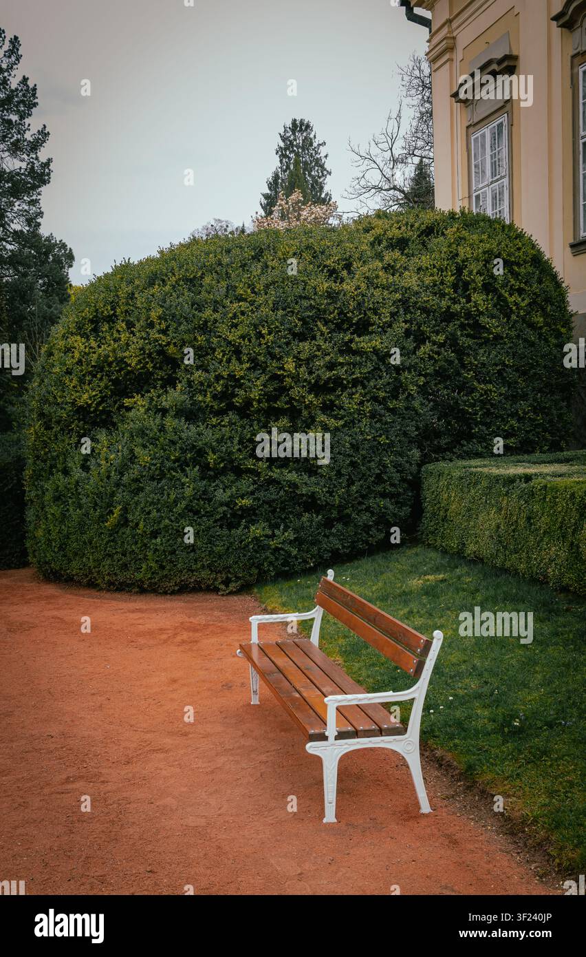 Ein ruhiger, vertikaler Schnappschuss einer leeren Holzbank, die auf einem roten Kiesweg in einem friedlichen Schlosspark steht. Stockfoto
