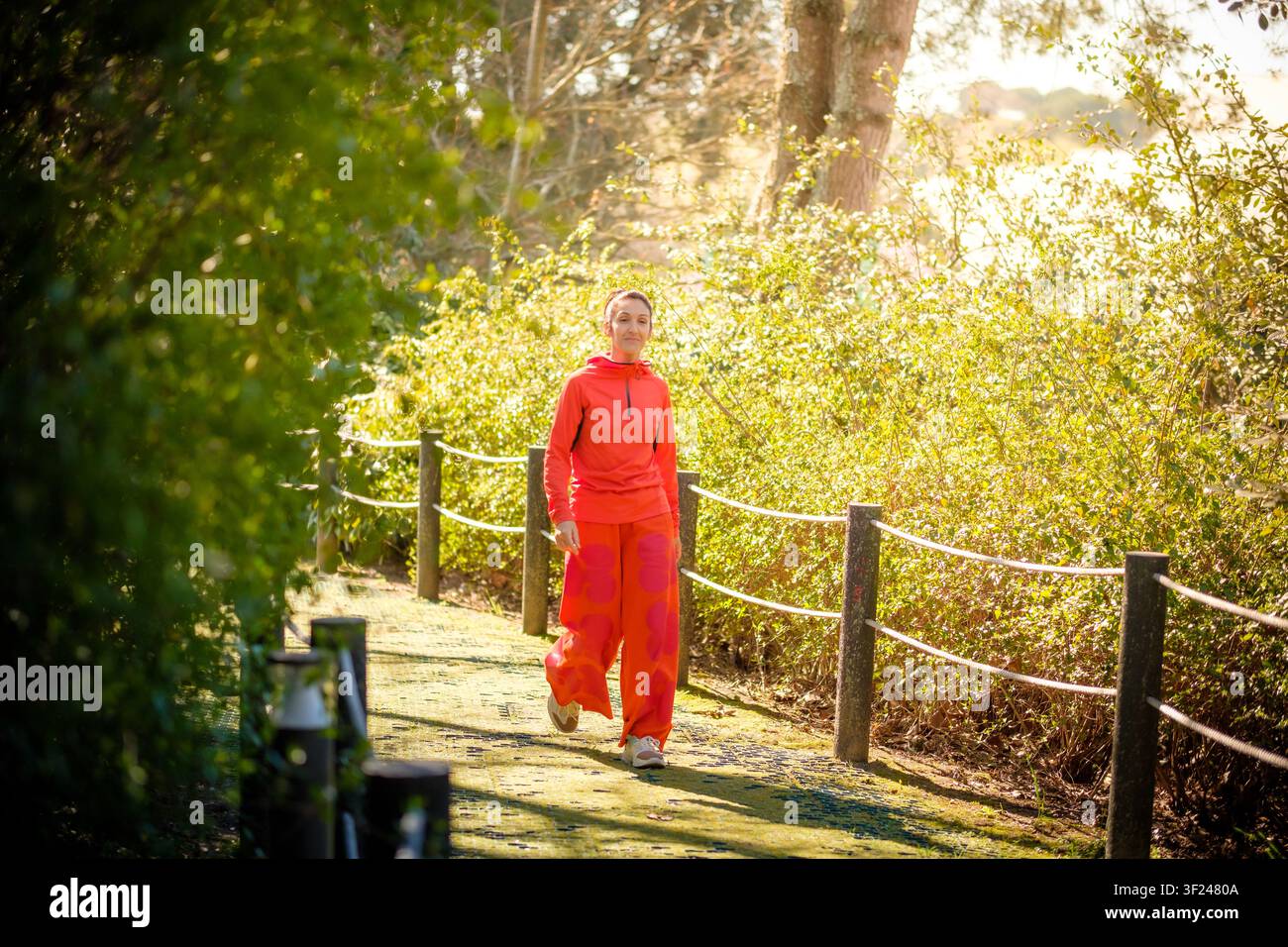 Frau in orangefarbener Sportkleidung geht auf einem Parkweg, umgeben von grünen Büschen und Sonnenlicht Stockfoto