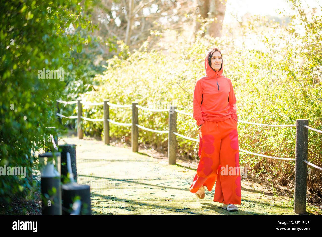 Frau, die einen orangefarbenen Kapuzenpullover und weite Hosen trägt, geht auf einem sonnigen Gartenweg Stockfoto