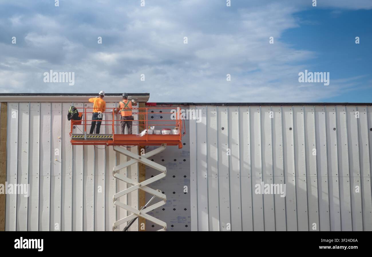 Zwei Bauarbeiter auf einem Scherenhubsteiger montieren isolierte Metallwandplatten an der Fassade eines Gewerbegebäudes unter einem dramatischen bewölkten Himmel. Indus Stockfoto