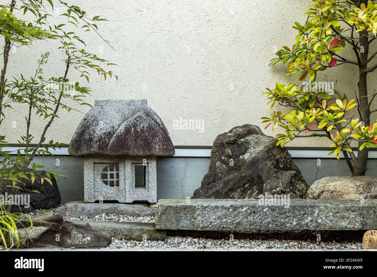 Traditionelle japanische Steinlaterne in einem friedlichen Zen-Garten mit natürlichen Steinen, grünem Laub und einer strukturierten Wand, die eine ruhige, minimalistische Szene schaffen Stockfoto