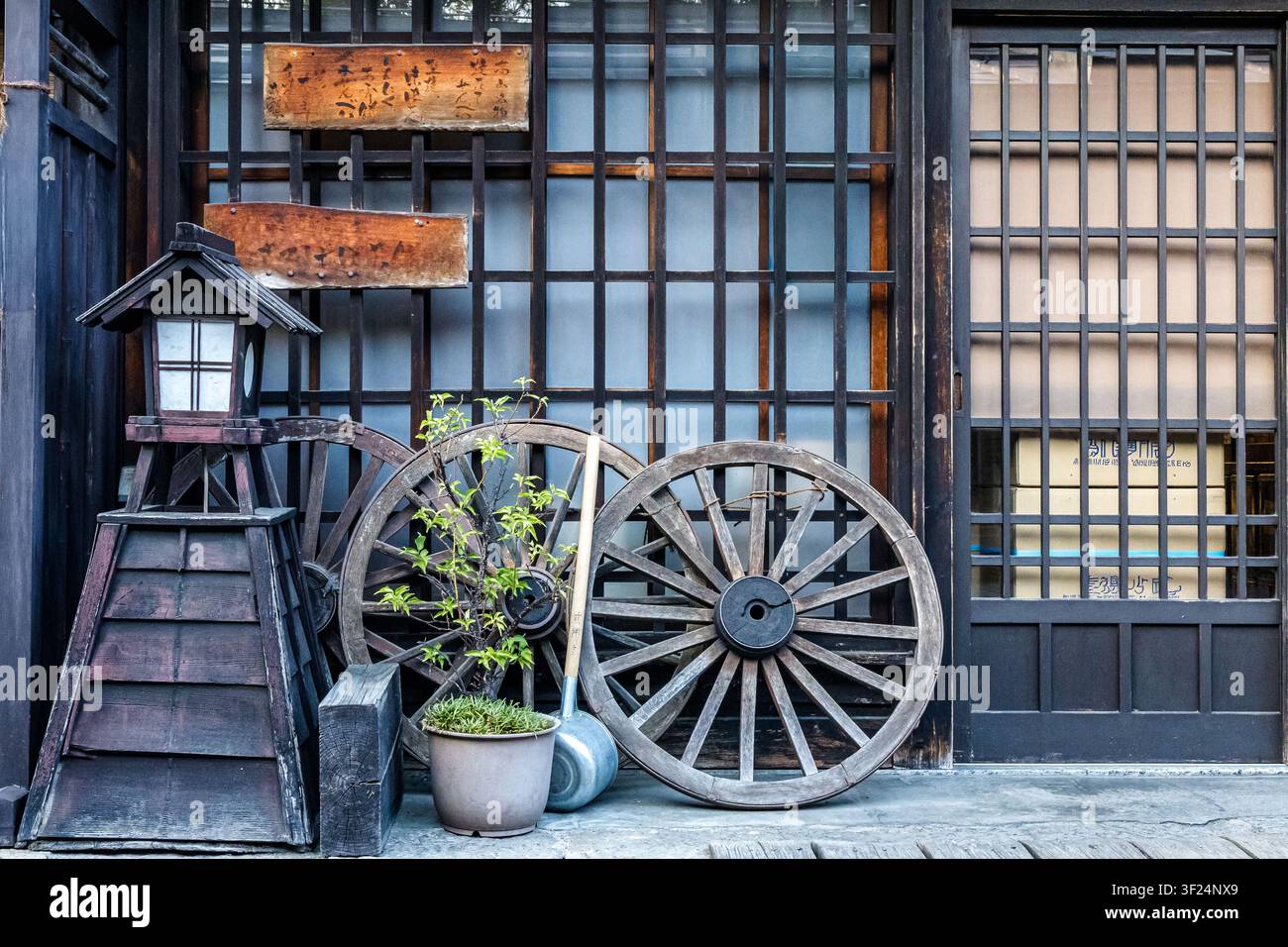 Traditionelle japanische Laterne in einem friedlichen Zen-Garten mit Wagenrädern, grünem Laub und einer strukturierten Wand, die eine ruhige, minimalistische Szene schaffen Stockfoto