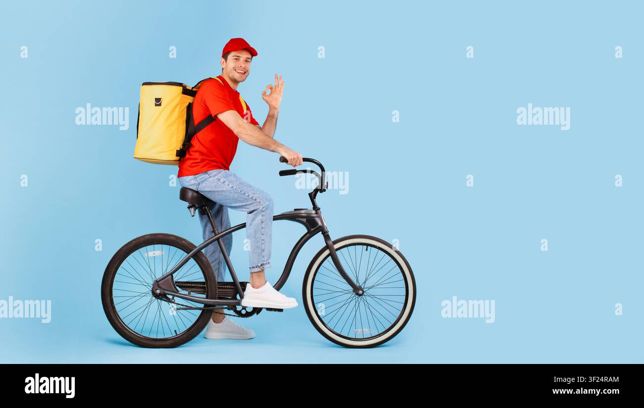 Lächelnder männlicher Lieferfahrer in rotem Hemd fährt Fahrrad, städtische Umgebung, Konzept für Lebensmittellieferung Stockfoto
