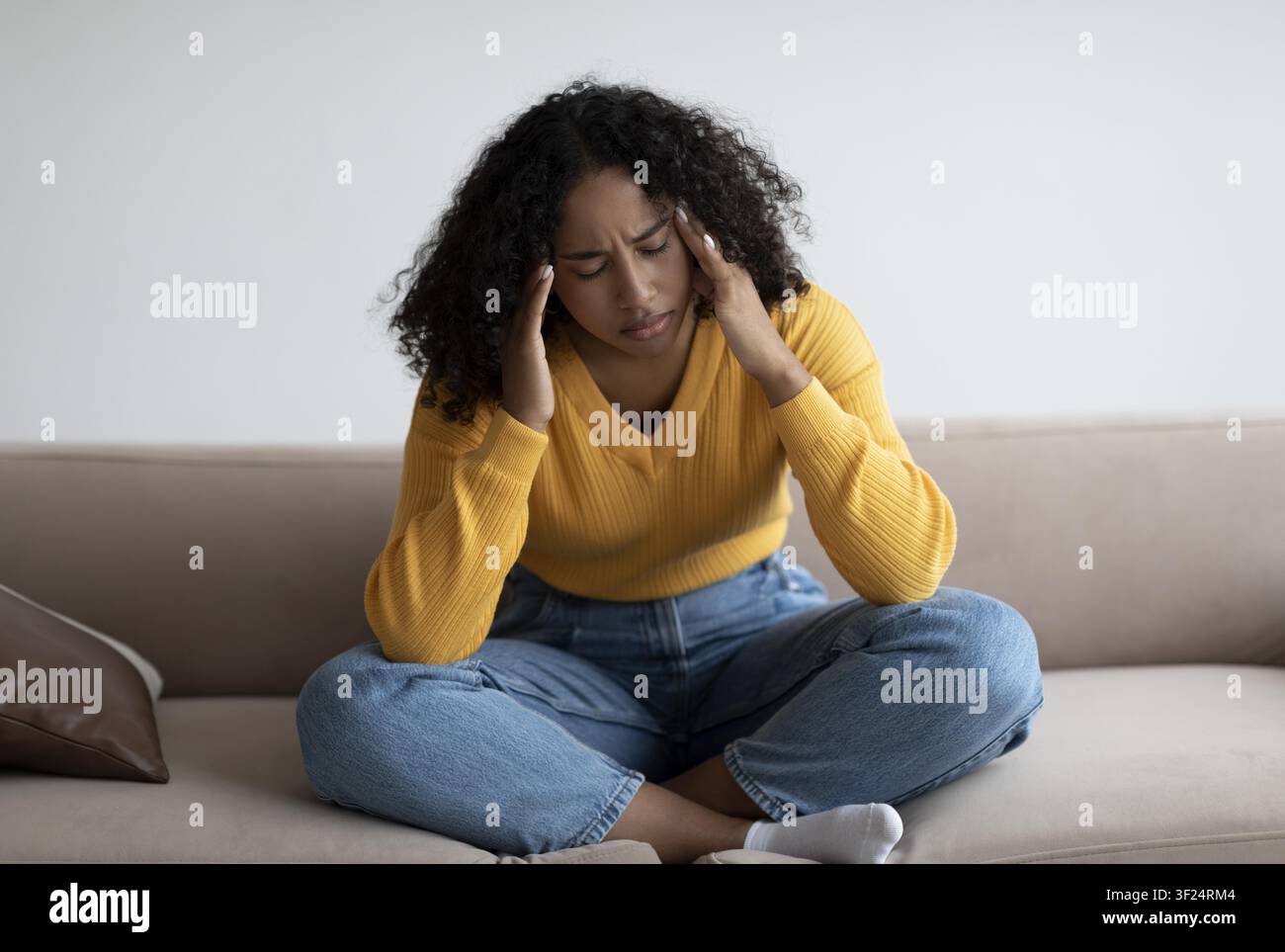Junge schwarze Frau mit Kopfschmerzen, die auf der Couch sitzt, häusliche Umgebung, Stresskonzept Stockfoto