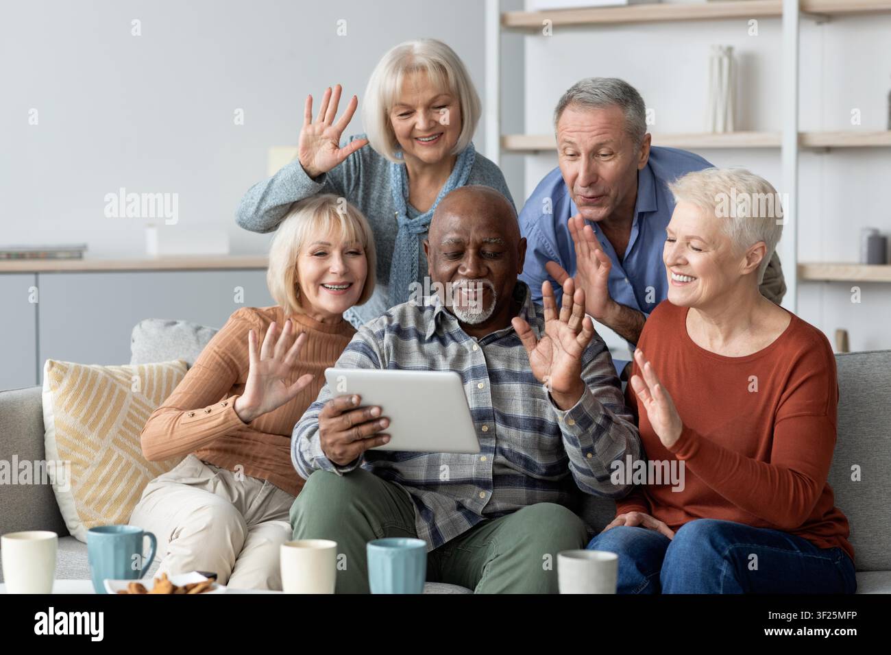 Fröhliche diverse Gruppe von Senioren, die mit einem Tablet auf der Couch interagieren, Heimsetting Stockfoto