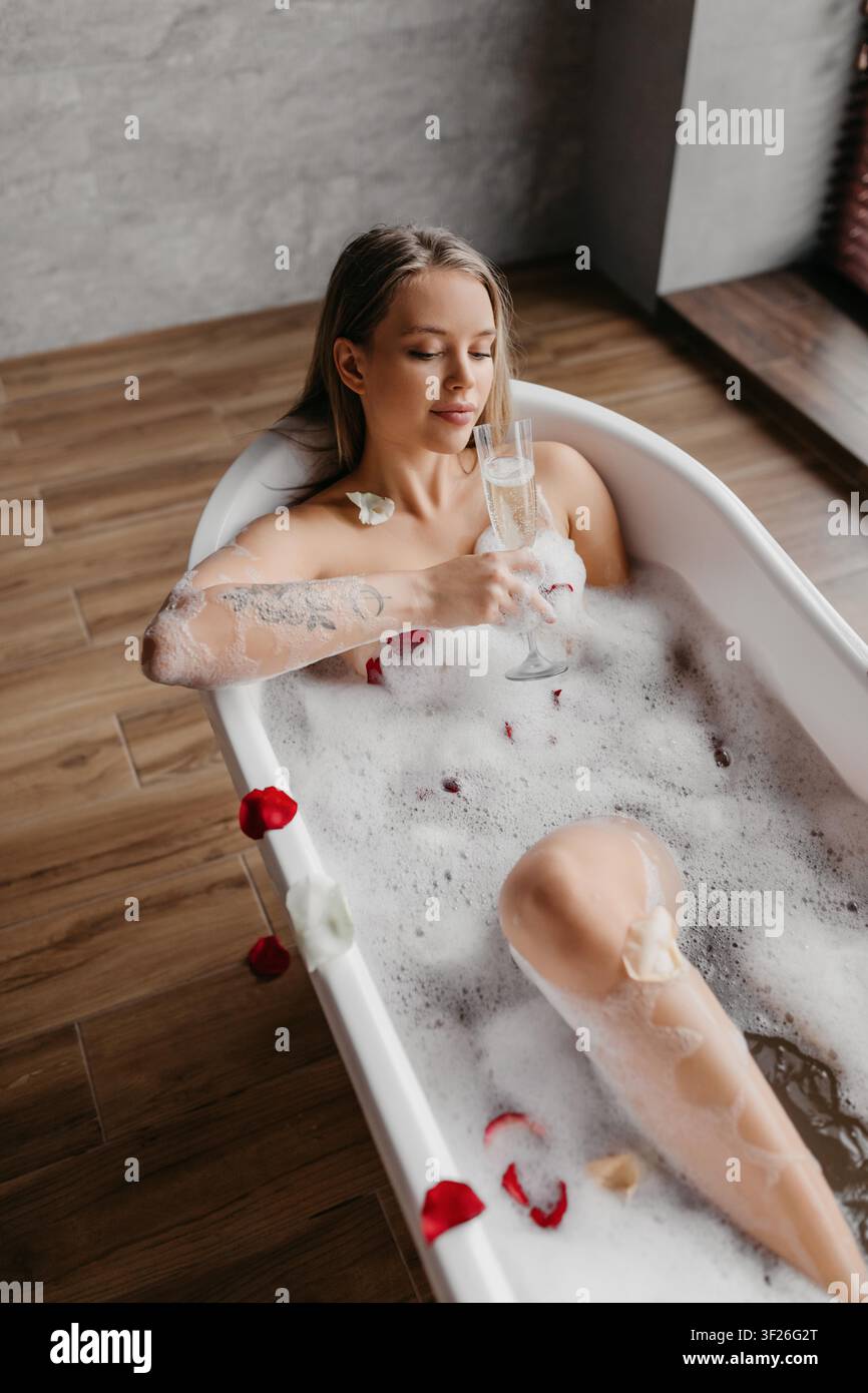 Junge Frau entspannt sich in der Badewanne mit Champagner und Rosenblättern, Spa-Konzept Stockfoto