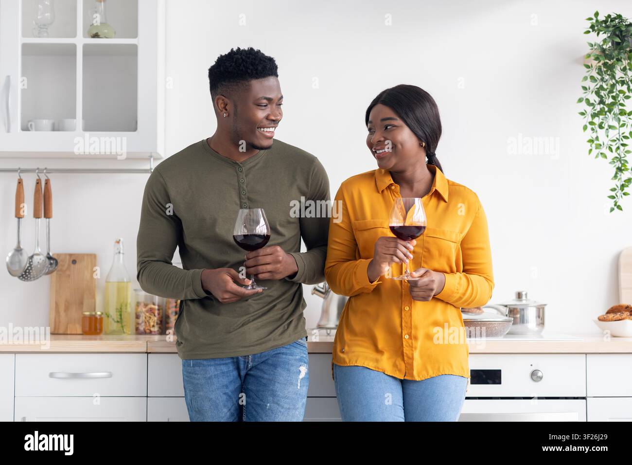 Fröhliches schwarzes Paar lächelt, während es Rotwein in einer modernen Küche trinkt, romantisches Abendkonzept Stockfoto