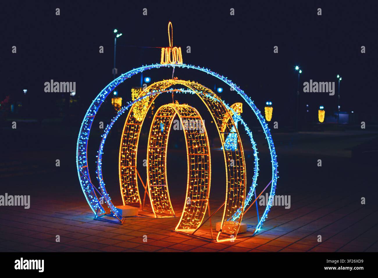 Leuchtende Ornamentskulptur leuchtet in der Nacht, goldene Bögen formen eine kugelförmige Kürbisform, umgeben von brillanten blauen LED-Ringen. Festliche Beleuchtung Stockfoto