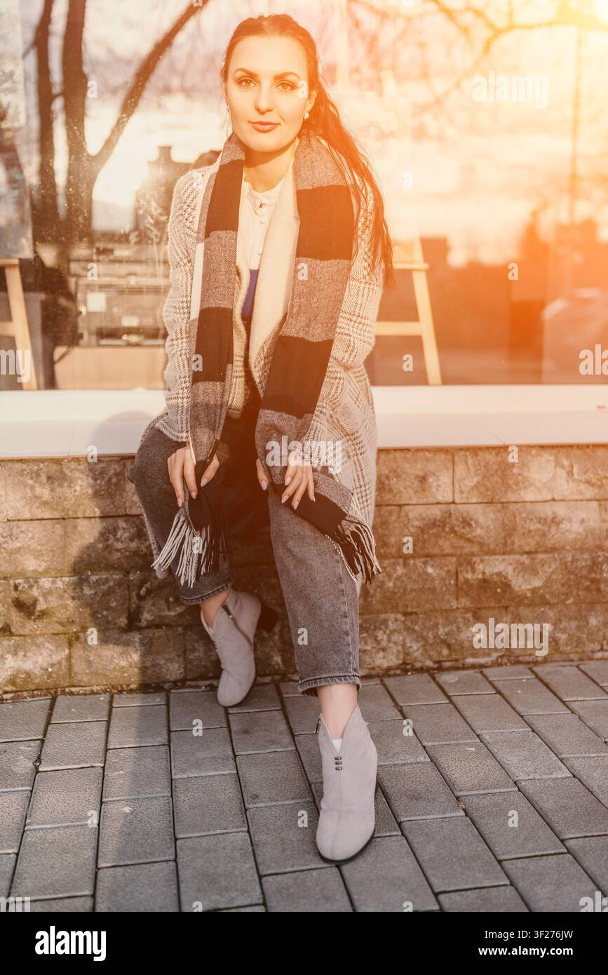 Damenschal Mode Junge Frau mit kariertem Schal und Mantel sitzt während der goldenen Stunde des Sonnenuntergangs auf einer Steinmauer Stockfoto