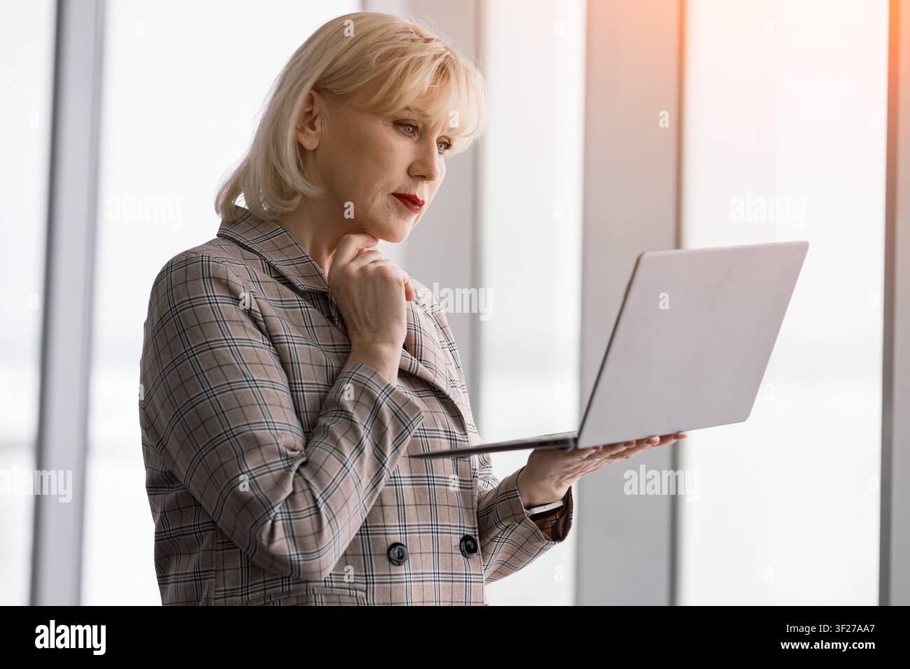 Senior-Beschäftigte steht mit einem Laptop in der Hand, arbeitet und konzentriert sich auf den Bildschirm, reflektiert mit nachdenklichem Ausdruck über Inhalte in einem modernen Stockfoto