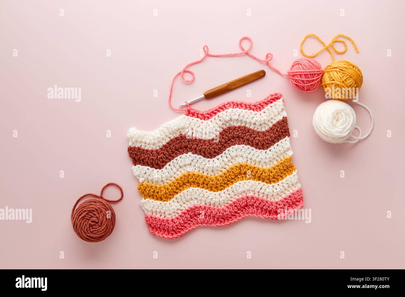 Farbenfrohes Häkelprojekt mit Wollknäulen und Häkelnadel auf pastellrosa Hintergrund. DIY-Häkelprojekt mit bunter Wolle und Häkelnadel, Flat Lay. Stockfoto