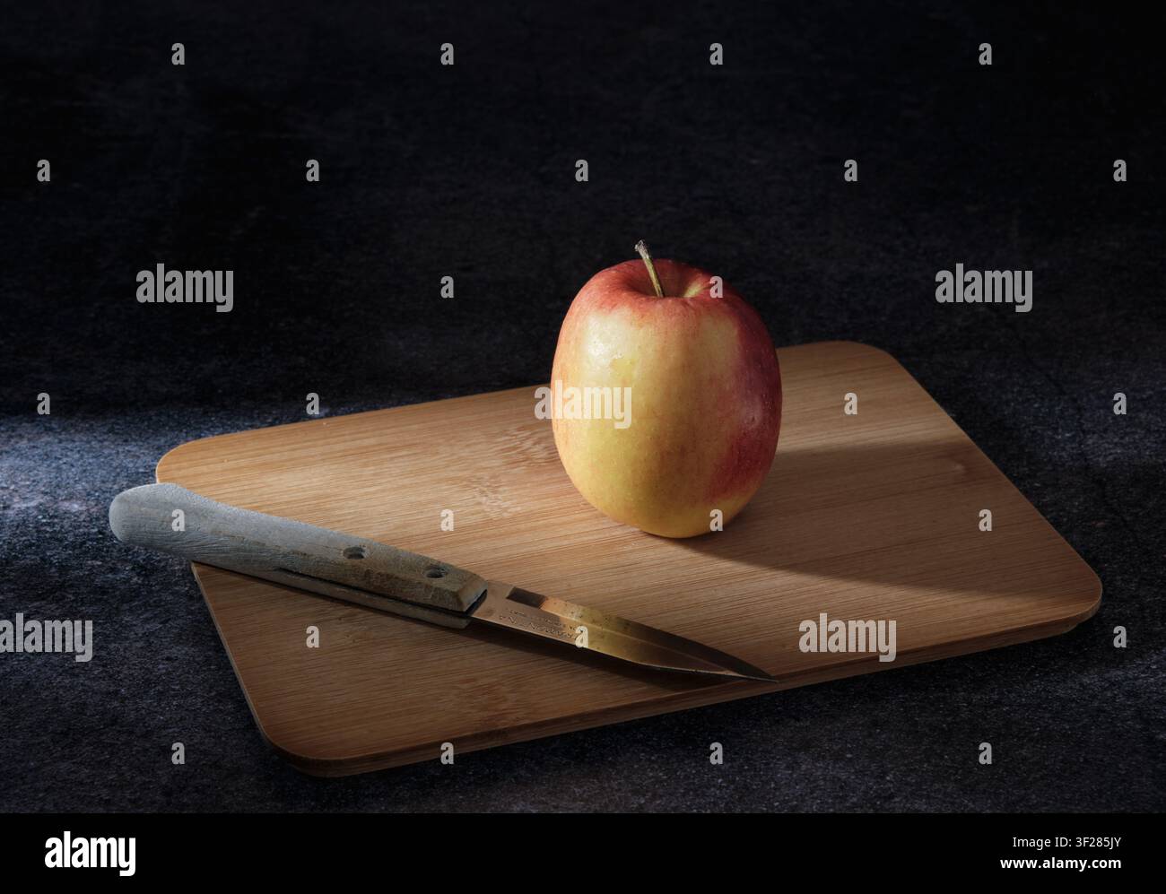 Apfel und rustikales Messer auf einem rustikalen Holzschneidebrett. Food-Fotografie im Fine-Art-Stil: Schwache Beleuchtung verleiht ein Fine-Art-, Altes-Meister-Gefühl. Stockfoto