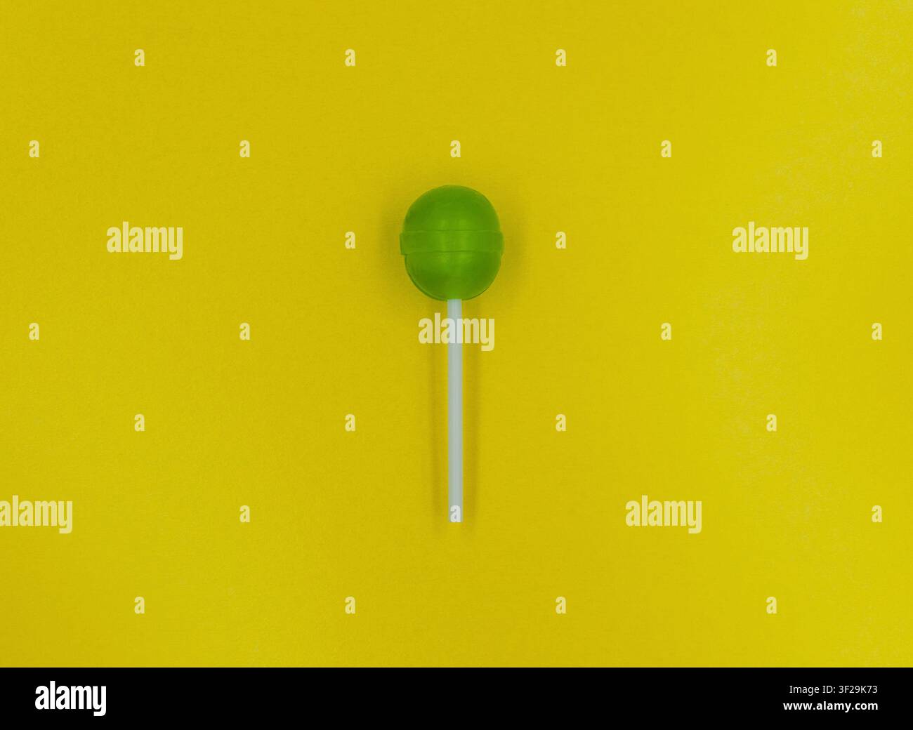Ein runder grüner harter Bonbon-Lutscher auf einem weißen Stiel steht in der Mitte vor einem leuchtend gelben Hintergrund. Stockfoto