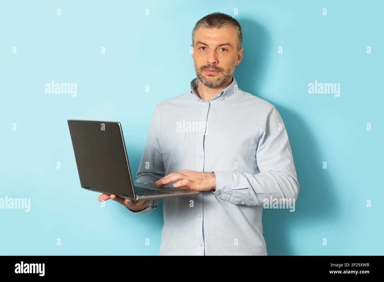 Ernster Kaukasier verwendet einen Laptop, steht vor hellblauer Kulisse, Technologiekonzept Stockfoto