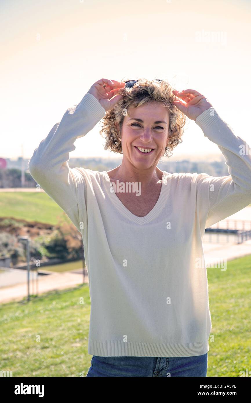 Frau lächelt und nimmt die Sonnenbrille ab und genießt einen Moment des Glücks im Park Stockfoto
