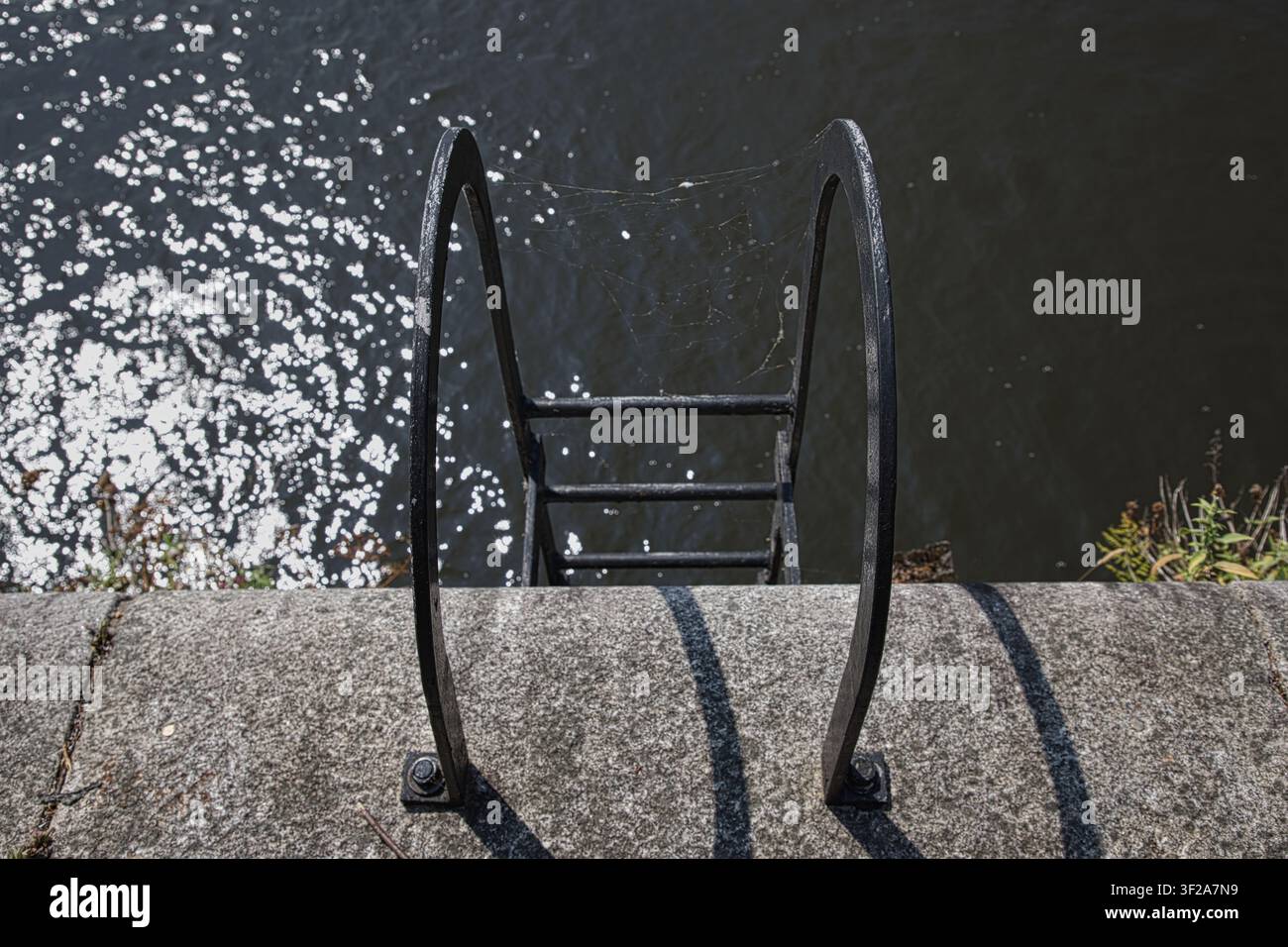 Eine Blick von oben auf eine schwarze Metall-Notleiter, die an einer Steinmole hinunter in funkelndes Kanalwasser führt, mit einem zarten Spinnennetz. Stockfoto