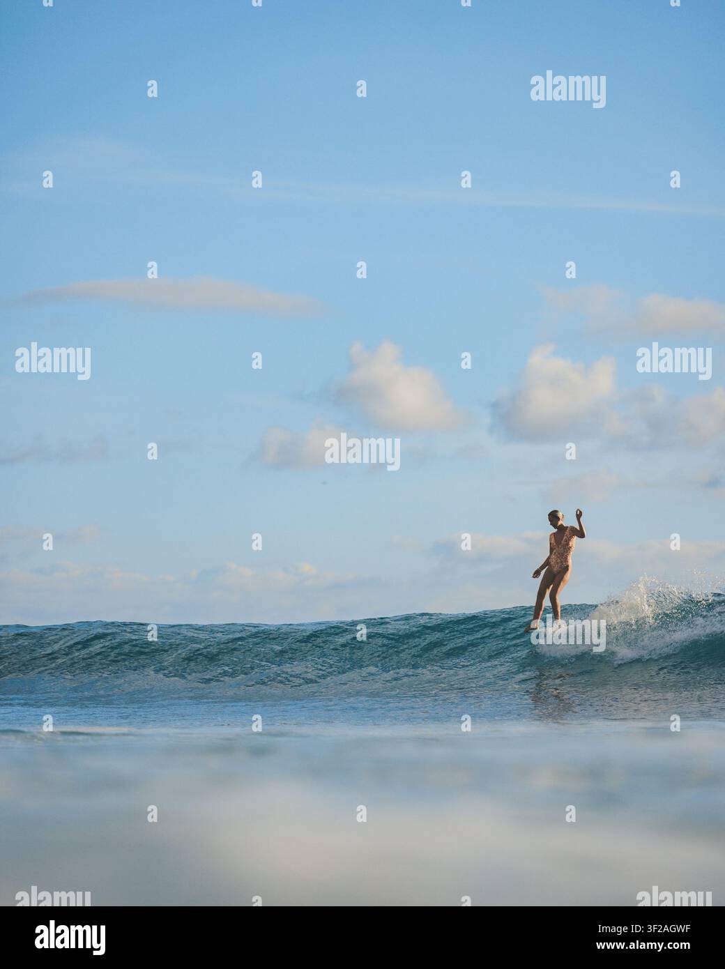 WAIKIKI, HONOLULU, HAWAII - 27. OKTOBER 2021: Ein einsamer Surfer steht auf einem Longboard und reitet eine kleine Welle unter einem weiten blauen Himmel, der mit Wolken übersät ist. Stockfoto