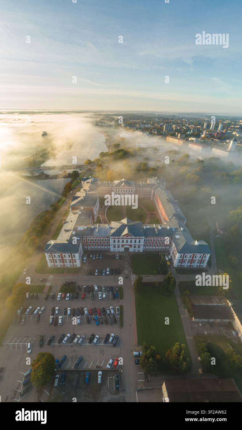 Luftaufnahme des Jelgava-Schlosses, umgeben von Morgennebel, üppigen Bäumen und einem Parkplatz in Jelgava, Lettland. Stockfoto