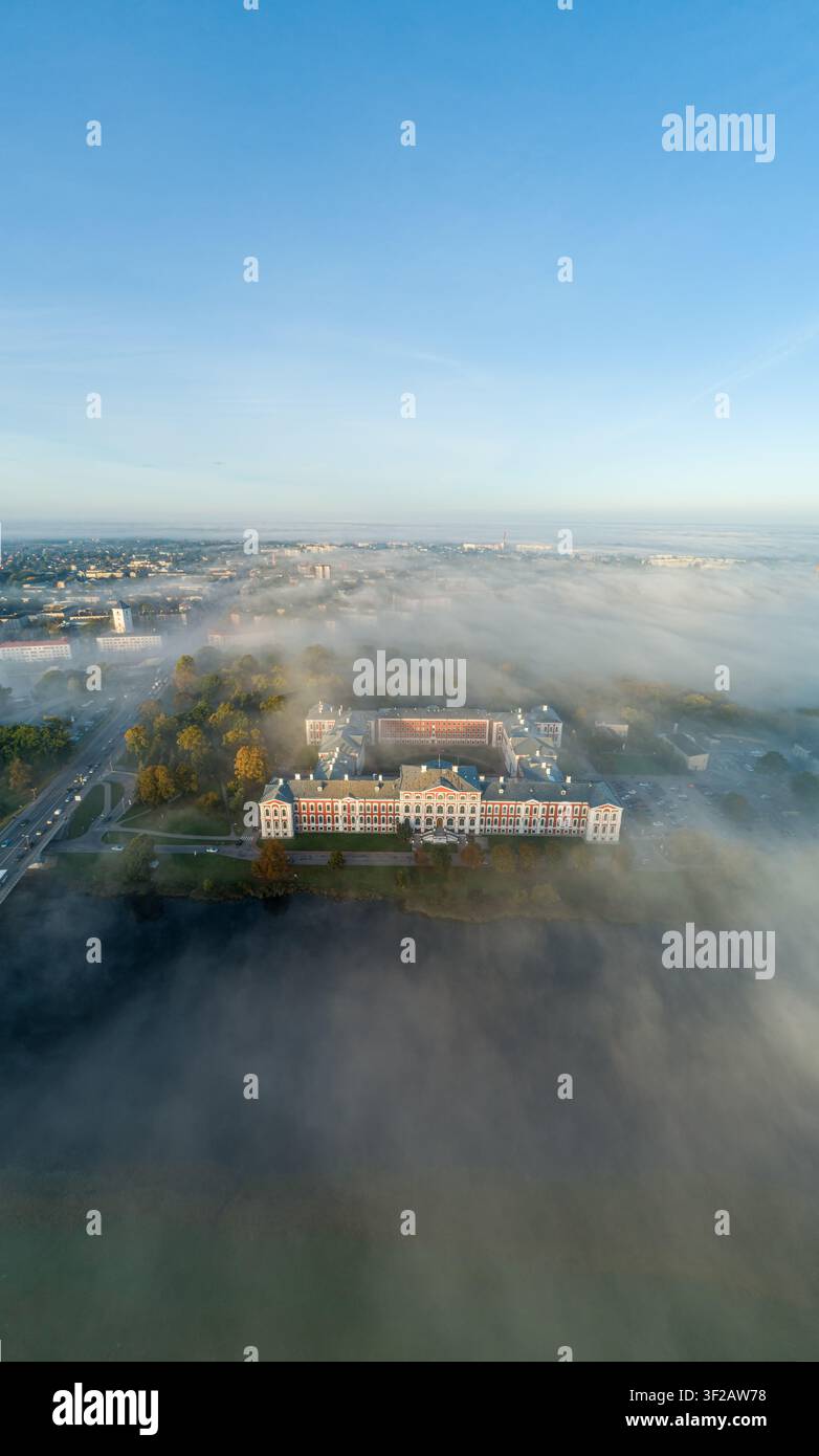 Luftbild des Jelgava-Schlosses, umgeben von dichtem Morgennebel, mit der Lielupe im Vordergrund und klarem blauem Himmel darüber in Jelgava, Lettland. Stockfoto