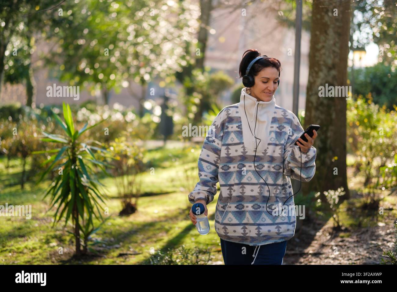 Frau genießt einen Spaziergang im Park, hört Musik mit Kopfhörern und überprüft ihr Smartphone Stockfoto