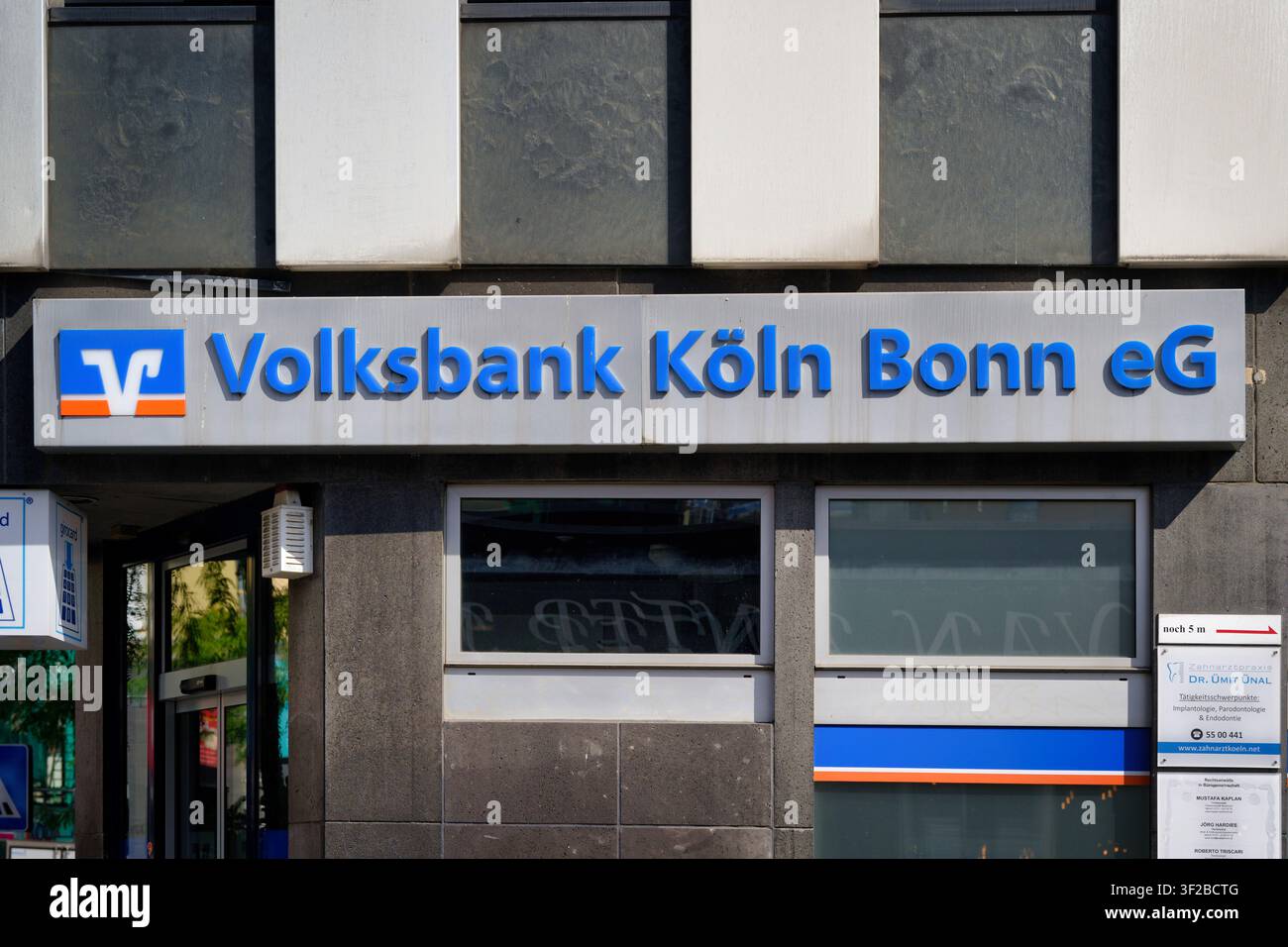 Köln, Deutschland, 15. Juli 2026: Offizielle Firmenbeschilderung und blaues Logo der Volksbank Köln Bonn eG Genossenschaftsbankfiliale an einem modernen Steingebäude Stockfoto
