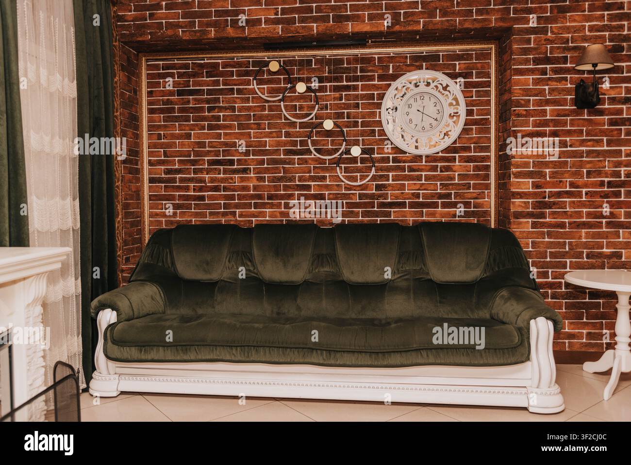 Elegantes Wohnzimmer-Interieur mit einem grünen Samtsofa, dekorativer Ziegelwand, Vintage-Uhr und warmer Umgebungsbeleuchtung. Gemütliches klassisches Wohndesign mit stilvollen Möbeln und luxuriöser Dekoration, perfekt für Innenarchitektur- und Immobilienkonzepte. Stockfoto
