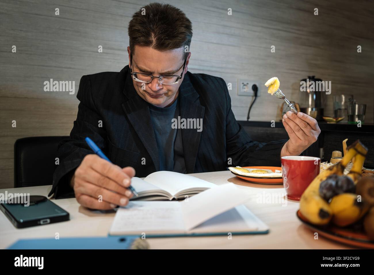 Konzentrierter Mann schreibt mit einem Stift in ein Notizbuch, während er während eines ruhigen Frühstücks zu Hause eine Gabel mit Essen hält. Stockfoto