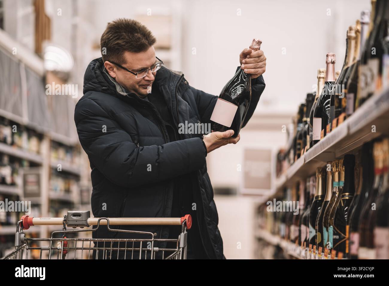 Zufriedener Kunde in blauer Daunenjacke betrachtet eine Flasche Sekt, während er im Alkoholgang eines Supermarkts steht. Stockfoto