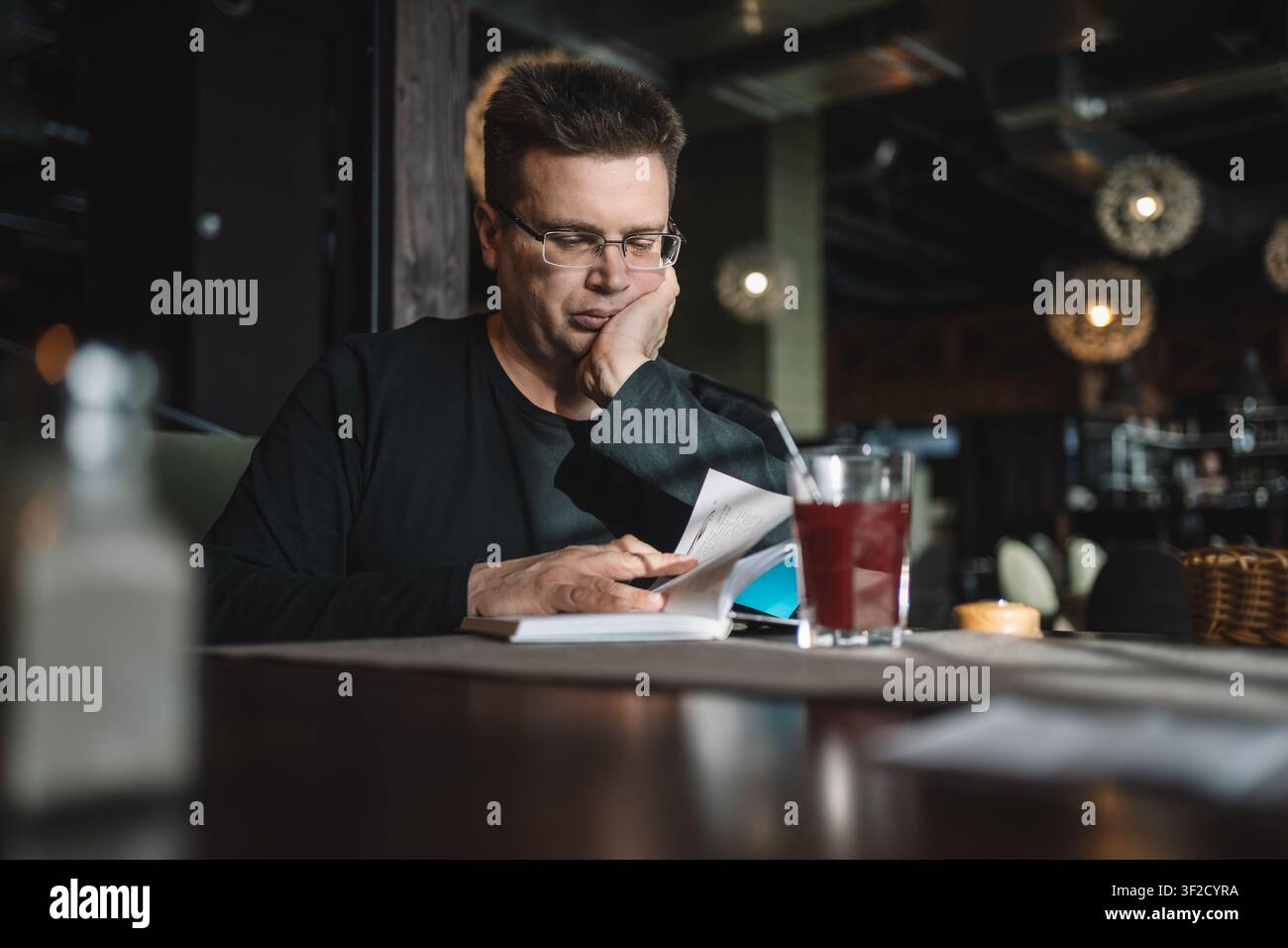 Mann mit Brille liest ein offenes Buch, während er sein Kinn auf die Hand stützt, mit einem Glas rotem Getränk auf dem Tisch. Stockfoto